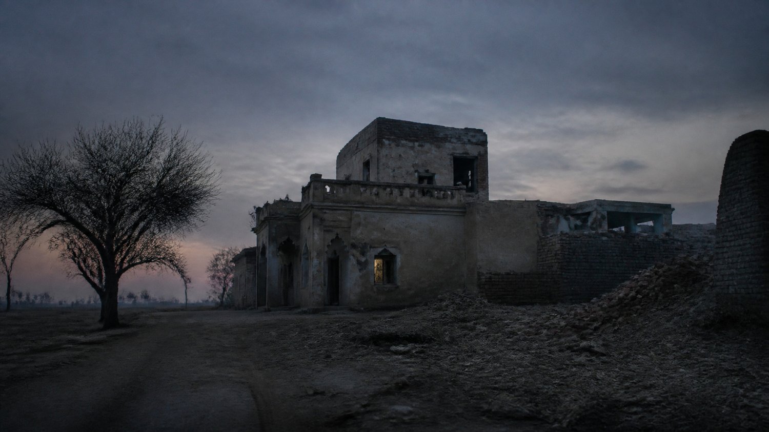 Rajjo Sadan (Exterior) — A crumbling ancestral village house off the GT Road — dust, dry fields, a siege at dusk. Where Satpal ends.